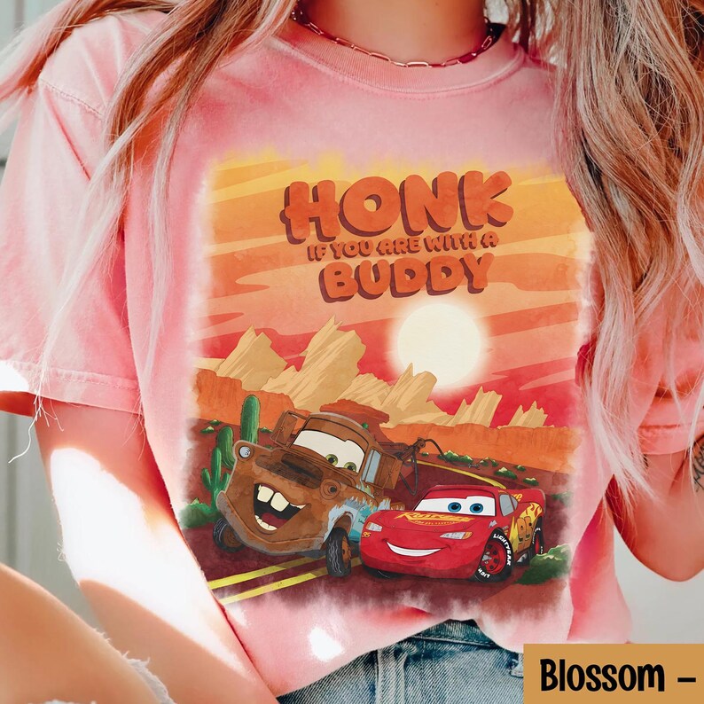 May include: A light pink t-shirt featuring a graphic of two cartoon cars, one brown and one red, with the text "HONK IF YOU ARE WITH A BUDDY". The background depicts a desert landscape with a setting sun.