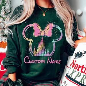 K&ouml;nnte beinhalten: Dunkelgr&uuml;nes Sweatshirt mit einem farbenfrohen Minnie-Mouse-Design, Schloss und Schleife. Das Design enth&auml;lt den Text "Custom Name" in Rosa. Das Sweatshirt ist ein Rundhals-Stil.