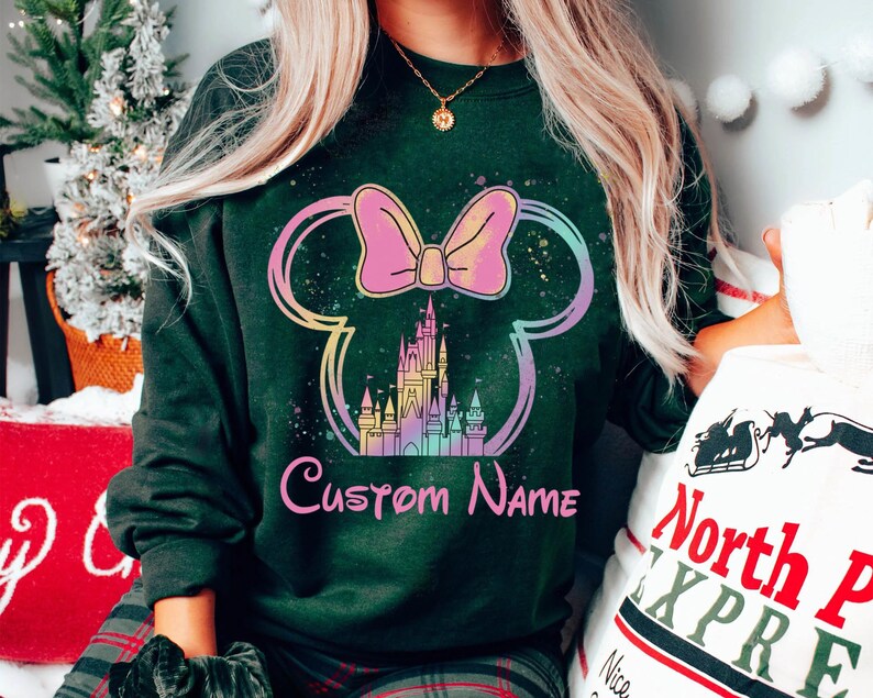 K&ouml;nnte beinhalten: Dunkelgr&uuml;nes Sweatshirt mit einem farbenfrohen Minnie-Mouse-Design, Schloss und Schleife. Das Design enth&auml;lt den Text "Custom Name" in Rosa. Das Sweatshirt ist ein Rundhals-Stil.