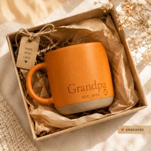 May include: Orange ceramic mug with "Grandpa" engraved, EST. 2026, and a watch icon. The mug has a gray base and is in a gift box with a tag that says "Made with Love".