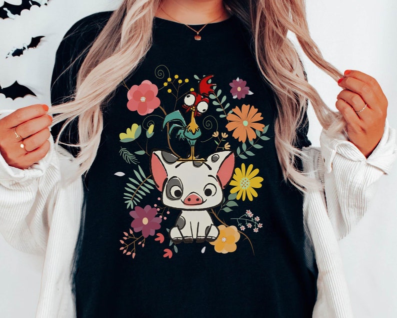 Pu&ograve; includere: Maglietta nera con un'immagine di un maiale e un gallo in stile cartone animato circondati da fiori colorati.