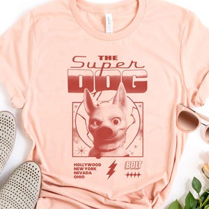 May include: A light peach-colored t-shirt featuring a graphic of a dog with the text "THE SUPER DOG." Below the dog graphic are the words "HOLLYWOOD NEW YORK NEVADA OHIO" and a lightning bolt. The shirt is paired with light-colored shoes and sunglasses.