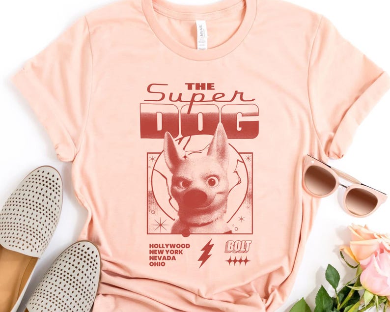 May include: A light peach-colored t-shirt featuring a graphic of a dog with the text "THE SUPER DOG." Below the dog graphic are the words "HOLLYWOOD NEW YORK NEVADA OHIO" and a lightning bolt. The shirt is paired with light-colored shoes and sunglasses.