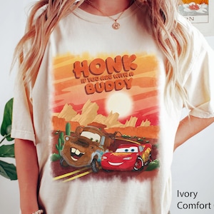 May include: Ivory-colored t-shirt featuring a graphic of two cartoon cars, Mater and Lightning McQueen, with a desert landscape backdrop. The text "HONK IF YOU ARE WITH A BUDDY" is displayed above the cars in orange letters.