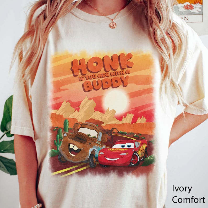 May include: Ivory-colored t-shirt featuring a graphic of two cartoon cars, Mater and Lightning McQueen, with a desert landscape backdrop. The text "HONK IF YOU ARE WITH A BUDDY" is displayed above the cars in orange letters.