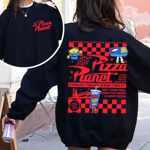 Op de afbeelding: Zwart sweatshirt met een rood-wit geruit patroon en een afbeelding van het Pizza Planet restaurant uit de Toy Story film. De afbeelding bevat de tekst "Pizza Planet", "Interstellar Fast-Food Fueling Station", "Friday Night Kids Night", en "Get 1 Free Pizza When You Order 2 Large Pizzas".
