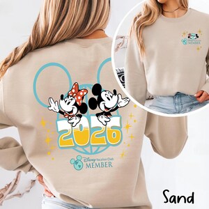 May include: Sand-coloured sweatshirt with Mickey and Minnie Mouse, the year "2026", and "Disney Vacation Club MEMBER". The design includes blue circles and yellow starbursts. A smaller version of the design is on the upper right.