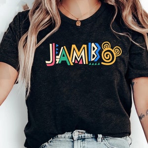 Puede incluir: Una camiseta negra con la palabra "JAMBO" en letras estilizadas de colores y un dise&ntilde;o de oreja de Mickey Mouse amarillo.