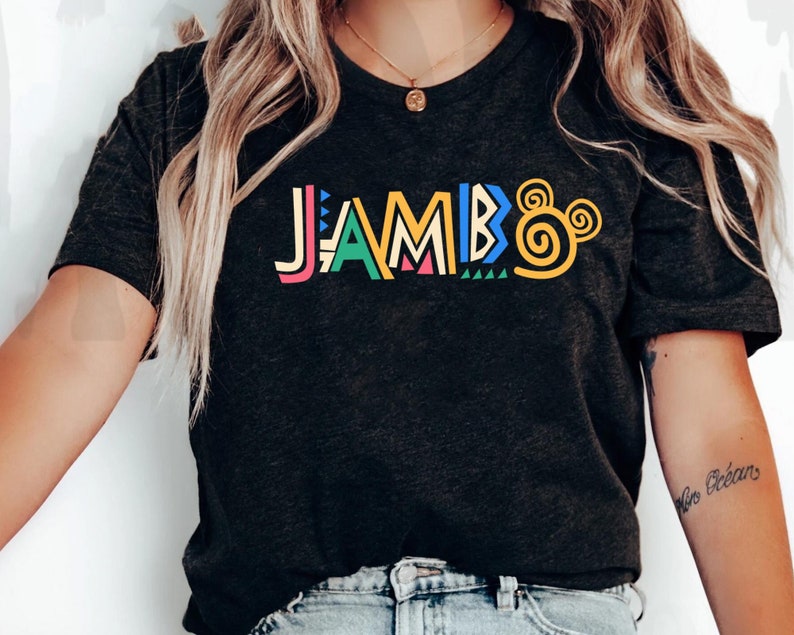 Puede incluir: Una camiseta negra con la palabra "JAMBO" en letras estilizadas de colores y un dise&ntilde;o de oreja de Mickey Mouse amarillo.