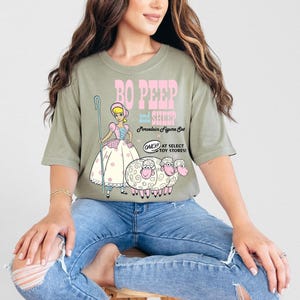 May include: A green t-shirt with a graphic of Bo Peep and her sheep from the Toy Story franchise. The text on the shirt reads "Bo Peep and Sheep Porcelain Figure Set Only at Select Toy Stores!"