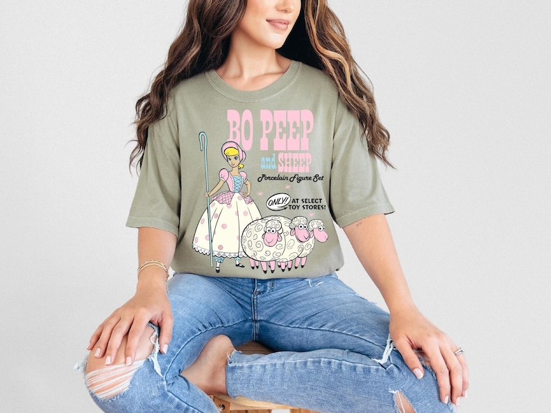 May include: A green t-shirt with a graphic of Bo Peep and her sheep from the Toy Story franchise. The text on the shirt reads "Bo Peep and Sheep Porcelain Figure Set Only at Select Toy Stores!"