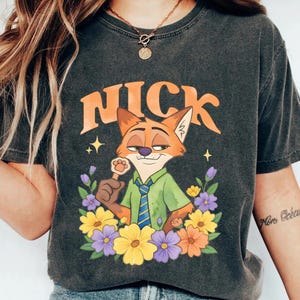 May include: Dark grey t-shirt with a cartoon fox character named Nick, surrounded by colourful flowers. The word "NICK" is printed above the fox in orange. The fox is wearing a green shirt and blue tie.