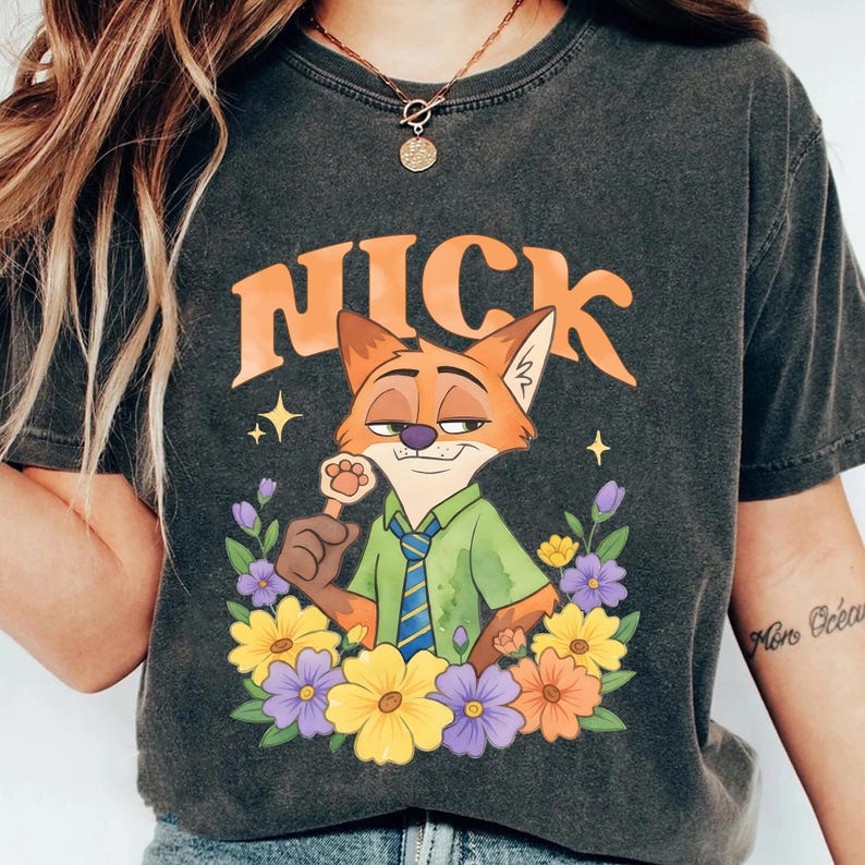 May include: Dark grey t-shirt with a cartoon fox character named Nick, surrounded by colourful flowers. The word "NICK" is printed above the fox in orange. The fox is wearing a green shirt and blue tie.