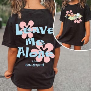 Puede incluir: Camiseta negra con la frase "Leave Me Alone" en texto azul claro, detalles de flores rosas y el logotipo de Disney Lilo & Stitch. Una imagen m&aacute;s peque&ntilde;a muestra un personaje de dibujos animados en la parte delantera.