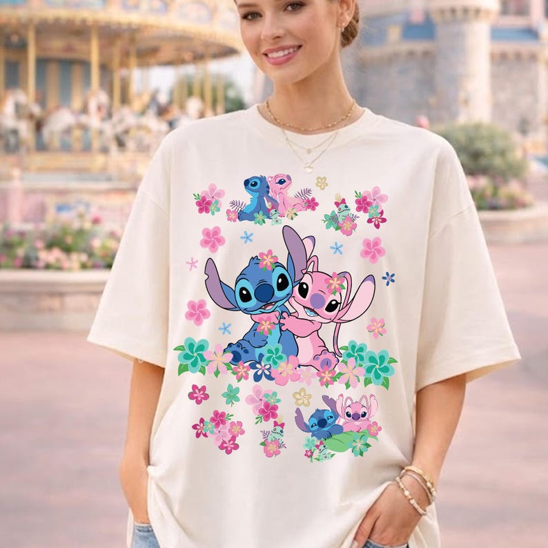 May include: Cream-colored t-shirt featuring a colorful graphic of the characters Stitch and Angel surrounded by pink and green flowers. The design includes multiple images of the characters. The shirt has short sleeves and a relaxed fit.