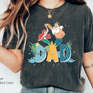 May include: Dark grey t-shirt with a colourful graphic of Ariel and King Triton dancing above the word "DAD" in a stylized font. The shirt has a vintage, faded look and the text "Pepper Comfort Color" is printed at the bottom.