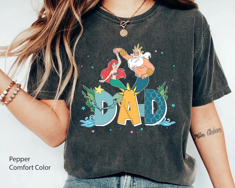 May include: Dark grey t-shirt with a colourful graphic of Ariel and King Triton dancing above the word "DAD" in a stylized font. The shirt has a vintage, faded look and the text "Pepper Comfort Color" is printed at the bottom.