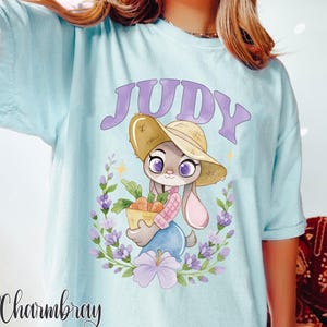 May include: Pale blue t-shirt with a cartoon bunny wearing a straw hat and carrying carrots. The name "JUDY" is above the bunny in purple. The design is framed by purple flowers and green foliage.