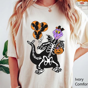 May include: An ivory t-shirt featuring a graphic of a purple dragon wearing a black skeleton costume. The dragon holds three orange balloons with black Mickey Mouse silhouettes and an orange jack-o'-lantern bucket. The text Ivory Comfort Color is visible.