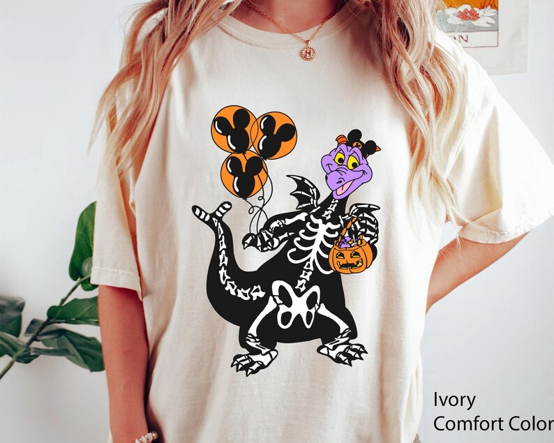 May include: An ivory t-shirt featuring a graphic of a purple dragon wearing a black skeleton costume. The dragon holds three orange balloons with black Mickey Mouse silhouettes and an orange jack-o'-lantern bucket. The text Ivory Comfort Color is visible.