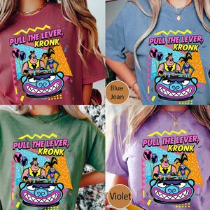 Puede incluir: Cuatro camisetas de diferentes colores con un dise&ntilde;o gr&aacute;fico retro que presenta un personaje de dibujos animados conduciendo un veh&iacute;culo con el texto "Pull the Lever, Kronk". Las camisetas son de color carmes&iacute;, azul vaquero, verde musgo y violeta.