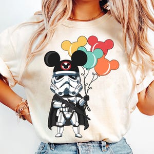 May include: A white Stormtrooper character wearing a black cape and Mickey Mouse ears, holding colorful balloons. The character is holding a blaster rifle in its right hand.