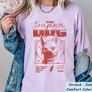 May include: Orchid-colored t-shirt with a red graphic of a dog and the text "THE SUPER DOG." Additional text includes "HOLLYWOOD NEW YORK NEVADA OHIO" and "BOLT." The shirt is a Comfort Color brand.