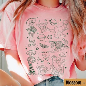 Puede incluir: Camiseta rosa claro con un dibujo lineal en blanco y negro de personajes de una pel&iacute;cula de animaci&oacute;n popular. La camiseta incluye el texto "Blossom -" en blanco.