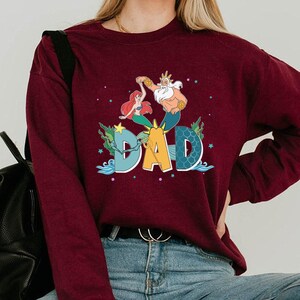 May include: A burgundy sweatshirt with a colourful graphic of Ariel, King Triton, and the word "DAD". The design includes aquatic elements like seaweed and stars, creating a whimsical, cartoon-like aesthetic.