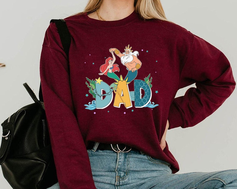 May include: A burgundy sweatshirt with a colourful graphic of Ariel, King Triton, and the word "DAD". The design includes aquatic elements like seaweed and stars, creating a whimsical, cartoon-like aesthetic.