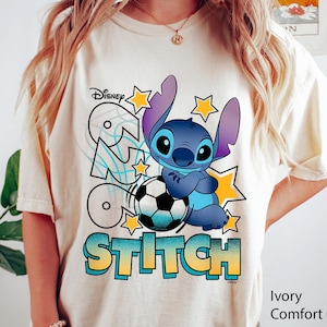 May include: Ivory-colored t-shirt featuring a cartoon image of Stitch from Disney's Lilo & Stitch, holding a soccer ball. The shirt has the word "Stitch" in a gradient blue and yellow font, with stars and the Disney logo.