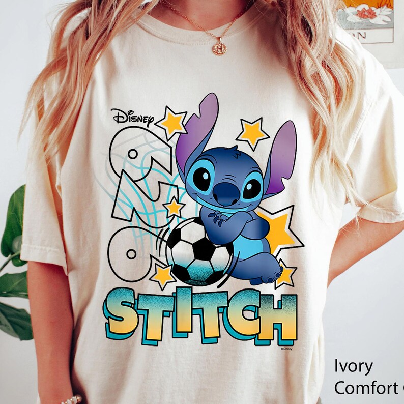 May include: Ivory-colored t-shirt featuring a cartoon image of Stitch from Disney's Lilo & Stitch, holding a soccer ball. The shirt has the word "Stitch" in a gradient blue and yellow font, with stars and the Disney logo.
