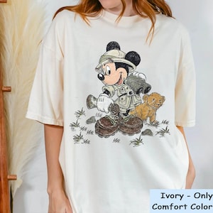 May include: A white t-shirt with a cartoon image of Mickey Mouse dressed as a safari explorer. Mickey is holding binoculars and walking with a small lion cub. The text "Ivory - Only For Comfort Color Shirt" is printed at the bottom of the shirt.
