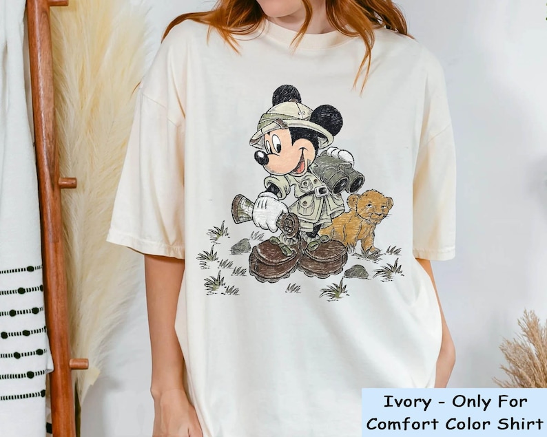 May include: A white t-shirt with a cartoon image of Mickey Mouse dressed as a safari explorer. Mickey is holding binoculars and walking with a small lion cub. The text "Ivory - Only For Comfort Color Shirt" is printed at the bottom of the shirt.