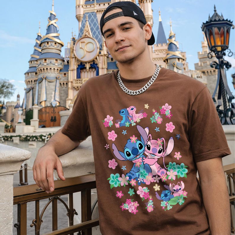 May include: Brown t-shirt featuring a colorful graphic of the characters Stitch and Angel surrounded by flowers. The shirt has short sleeves and a crew neck. The background includes a castle and a person wearing a black cap and silver chain.