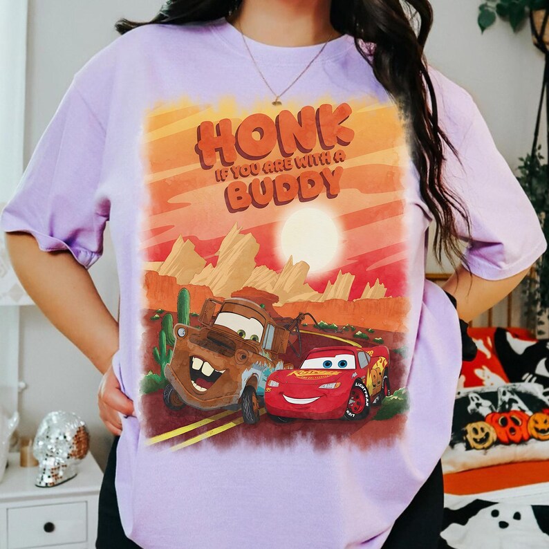 May include: A light purple t-shirt featuring a graphic of the characters Mater and Lightning McQueen from the movie Cars. The graphic includes the text "HONK IF YOU ARE WITH A BUDDY" in orange and brown.