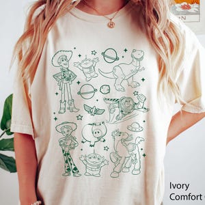 Puede incluir: Camiseta Comfort Colors color crema con un dise&ntilde;o en verde de personajes de una popular pel&iacute;cula de animaci&oacute;n. El dise&ntilde;o incluye un vaquero, un dinosaurio, un guardi&aacute;n espacial y otras figuras reconocibles. La camiseta tiene las palabras "Ivory Comfort" impresas en la parte inferior derecha.