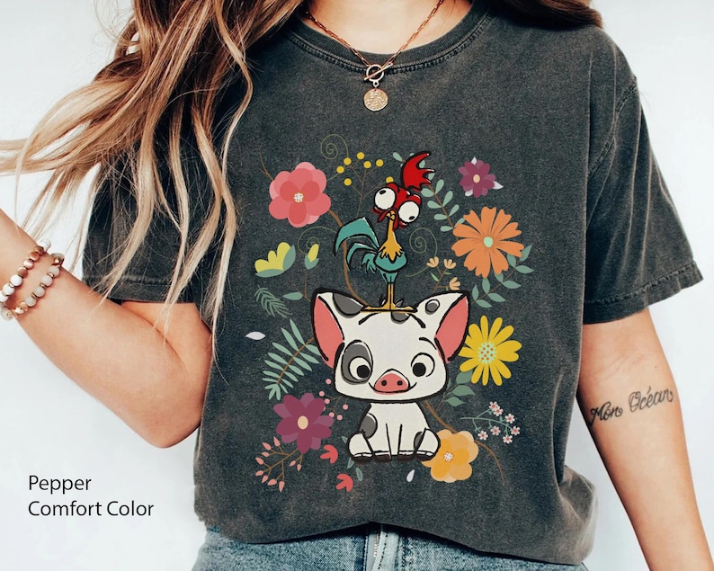 Pu&ograve; includere: Una maglietta grigia scura con un'immagine di un maiale e un gallo in stile cartone animato con fiori e foglie. Il testo "Moana Ocean" &egrave; sulla manica destra.