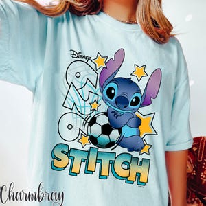 May include: Light blue t-shirt featuring the Disney character Stitch with a soccer ball. The design includes the word "Stitch" in a gradient font, along with stars and the Disney logo. The shirt has short sleeves.