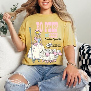 May include: A yellow t-shirt with a graphic of Bo Peep and her sheep from the Toy Story movie. The text on the shirt reads "Bo Peep and her Sheep Porcelain Figure Set. Only! At Select Toy Stores!"