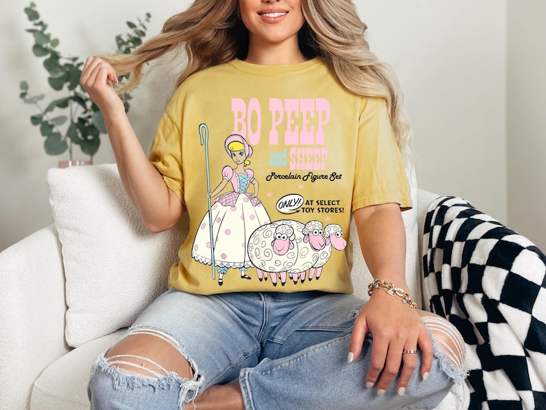 May include: A yellow t-shirt with a graphic of Bo Peep and her sheep from the Toy Story movie. The text on the shirt reads "Bo Peep and her Sheep Porcelain Figure Set. Only! At Select Toy Stores!"