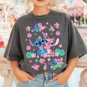 May include: Dark gray t-shirt featuring a graphic of the characters Stitch and Angel embracing, surrounded by colorful flowers. The design includes additional images of the characters and floral accents. The shirt is a relaxed fit and has short sleeves.