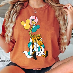 May include: A brown t-shirt with a cartoon lion wearing a crown and holding balloons. The lion is wearing a blue and white robe. The balloons are yellow, blue, and pink.