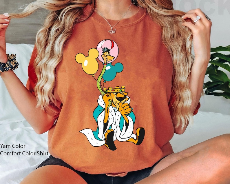 May include: A brown t-shirt with a cartoon lion wearing a crown and holding balloons. The lion is wearing a blue and white robe. The balloons are yellow, blue, and pink.