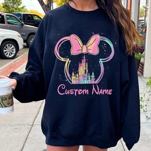 K&ouml;nnte beinhalten: Schwarzes Sweatshirt mit einem farbenfrohen Design aus Schleife, Mickey Mouse Ohren und Schloss. Die Worte "Custom Name" sind darunter in rosa Schrift gedruckt. Das Design hat einen Aquarell-Effekt.