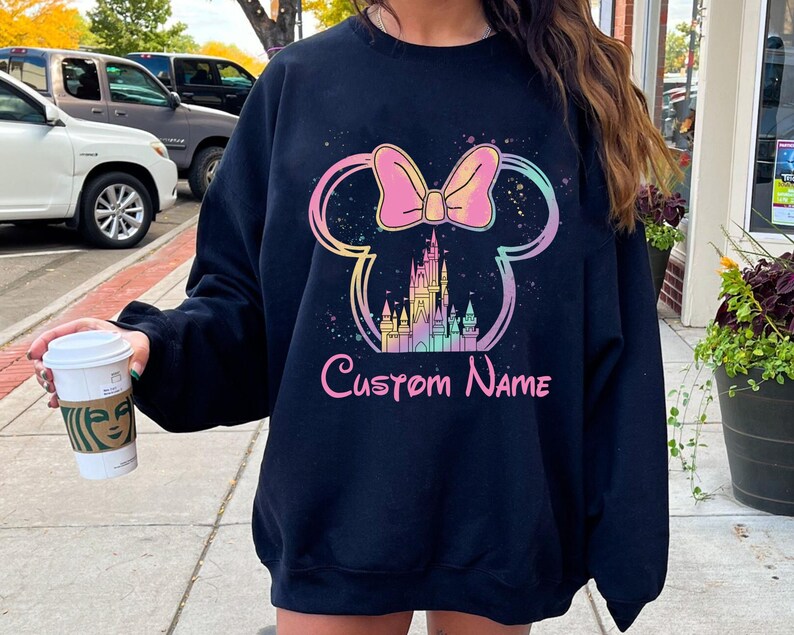 K&ouml;nnte beinhalten: Schwarzes Sweatshirt mit einem farbenfrohen Design aus Schleife, Mickey Mouse Ohren und Schloss. Die Worte "Custom Name" sind darunter in rosa Schrift gedruckt. Das Design hat einen Aquarell-Effekt.