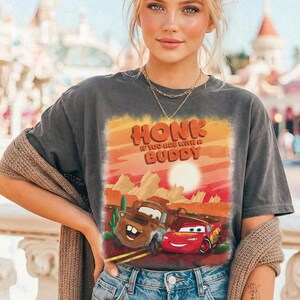 May include: Dark gray t-shirt featuring a graphic of two cartoon cars with the text "HONK IF YOU ARE WITH A BUDDY". The design includes a desert landscape with a sun and cacti. The shirt is worn by a person.