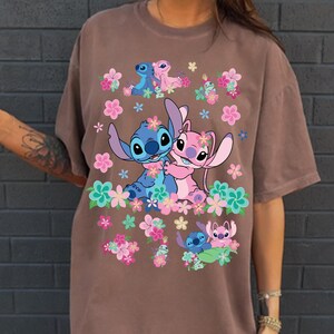 May include: Brown t-shirt featuring a colorful graphic of the characters Stitch and Angel from Lilo & Stitch, surrounded by pink, blue, and green flowers. The design includes multiple images of the characters in various poses.