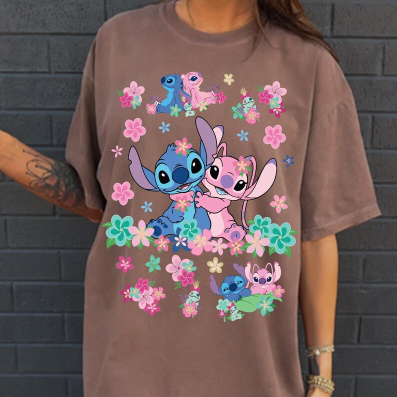 May include: Brown t-shirt featuring a colorful graphic of the characters Stitch and Angel from Lilo & Stitch, surrounded by pink, blue, and green flowers. The design includes multiple images of the characters in various poses.