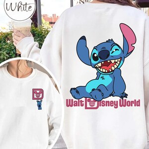 May include: White sweatshirt featuring a blue cartoon character with large ears and a wide grin. The character is sitting and winking. The text "Walt Disney World" is below the character. A smaller version of the character is on the front.